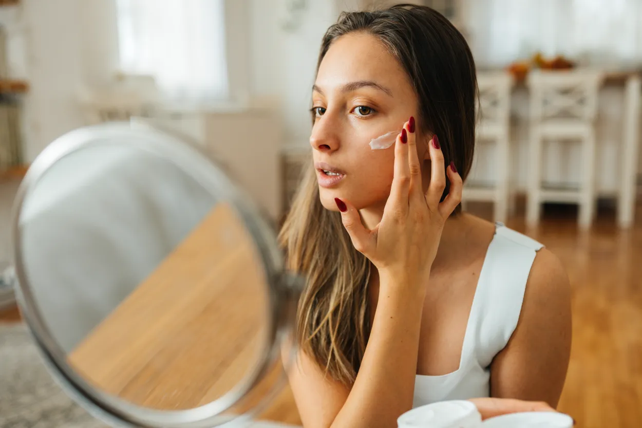 Foto de mujer frente al espejo aplicándose crema en la cara.