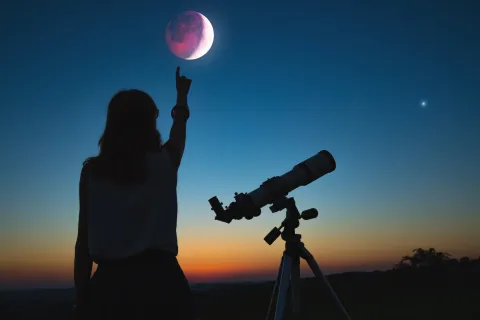 Una mujer con un telescopio mirando una luna eclipsada.